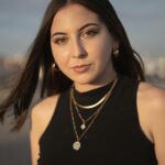 Portrait of a young woman with jewelry, black crop top, and windblown hair at sunset.