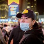 A young adult in Times Square at night wearing a mask and hat, capturing urban nightlife.